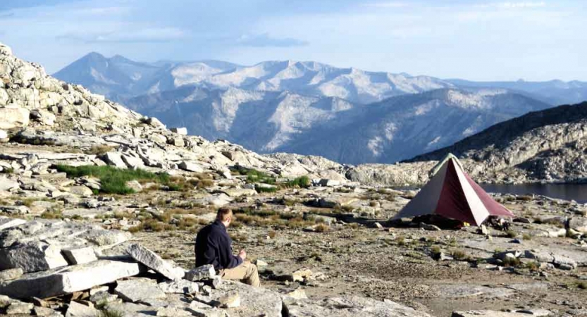 A person sits on a rock a few feet away from a tent. Behind the tent is a vast mountainous landscape.